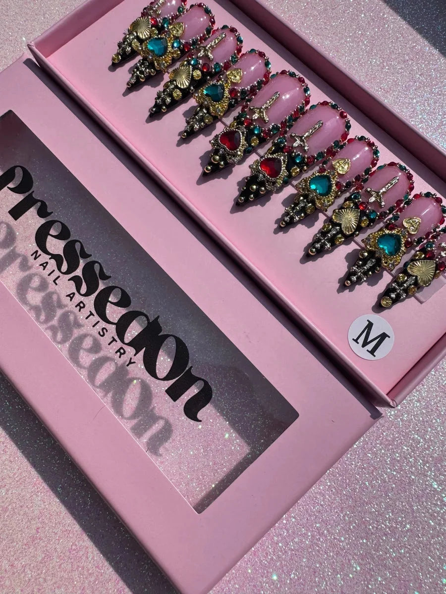 Set of medium stiletto press-on nails featuring pink base color with ornate embellishments, rhinestones, and jewels in red, green, and gold tones. Decorated with heart and cross charms and black pointed tips.