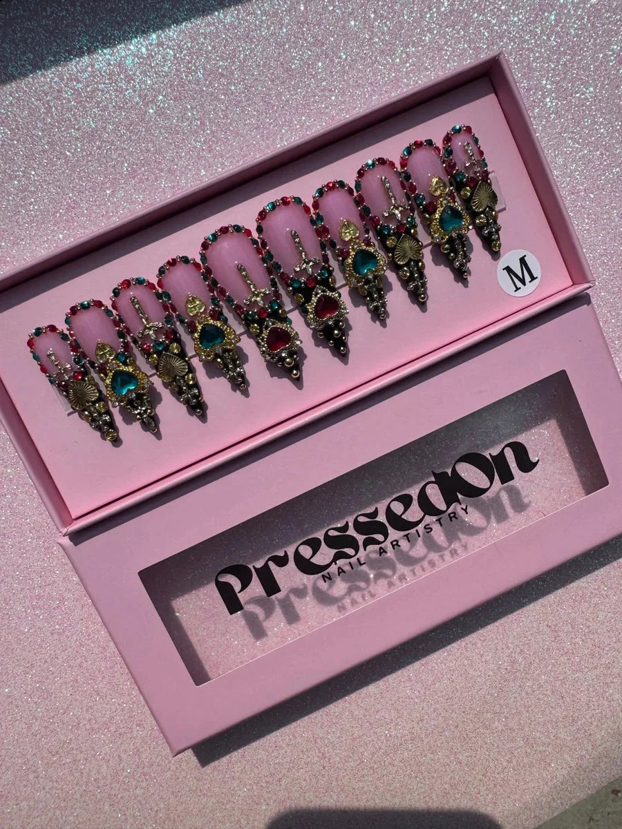 Medium stiletto press-on nails with a pink base, heavily decorated with red, teal, and gold jewels, hearts, crosses, and rhinestones. The nails have black accents for a unique, blinged-out style.