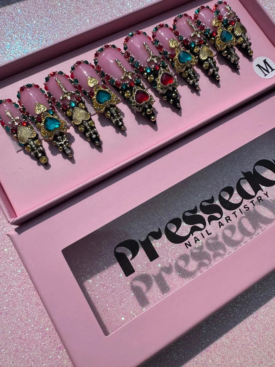 Medium stiletto press-on nails featuring a pink base, rhinestones, colorful jewels, gold hearts and crosses, and black tips. These blinged-out nails feature red, green, and gold embellishments.