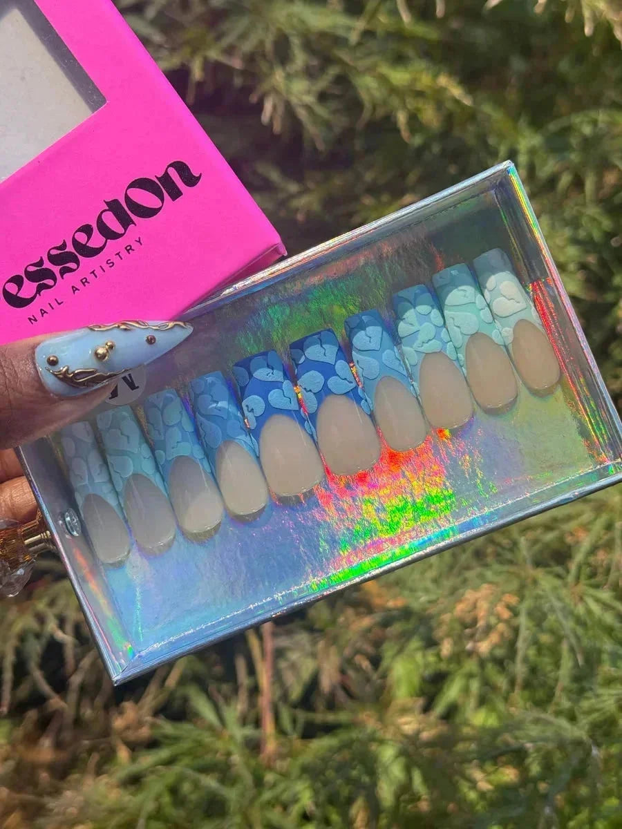 Short press-on nails feature a light nude base with a blue floral design and dark blue French tips. The nails have a rounded shape.