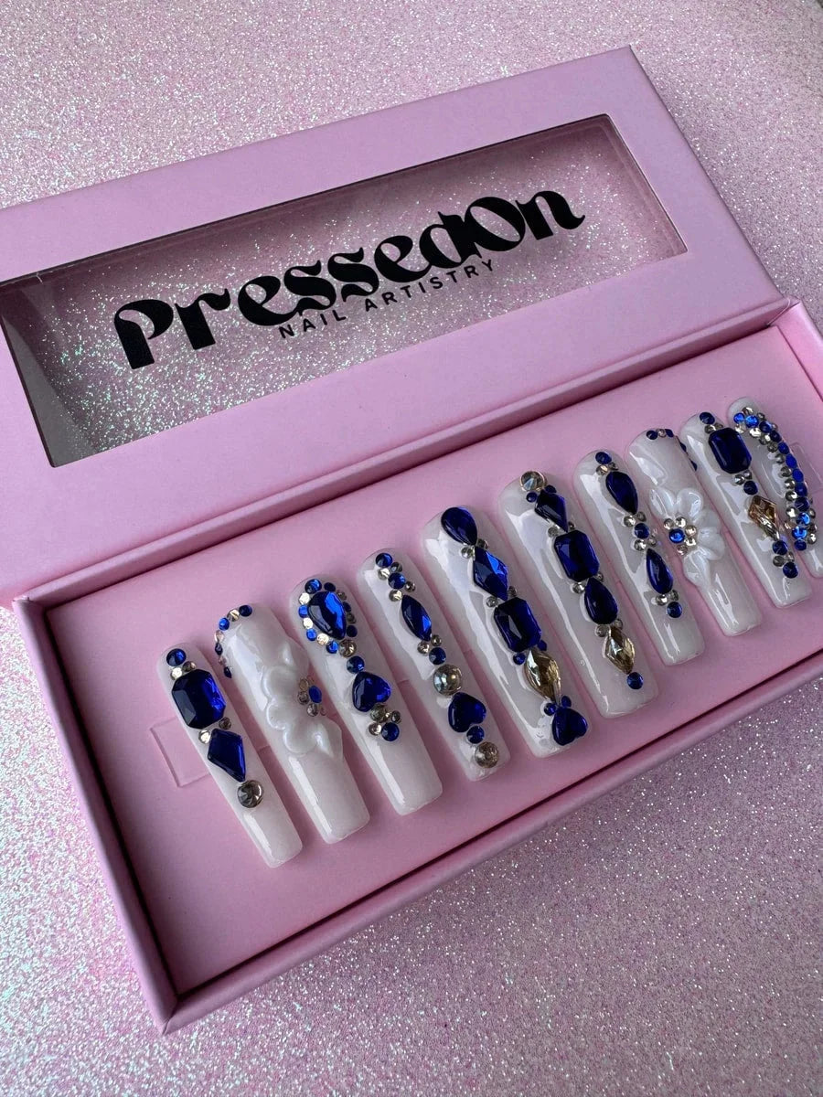 Long white coffin press-on nails adorned with blue diamond rhinestones and gems, as well as silver accents. Several nails have a 3D white flower charm.