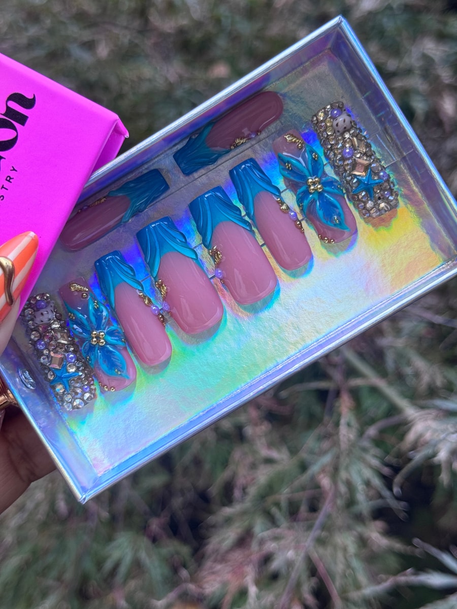 These are medium length pink and turquoise blue press-on nails with a tide design, featuring gold beads, rhinestones, and one fully rhinestone-covered accent nail.