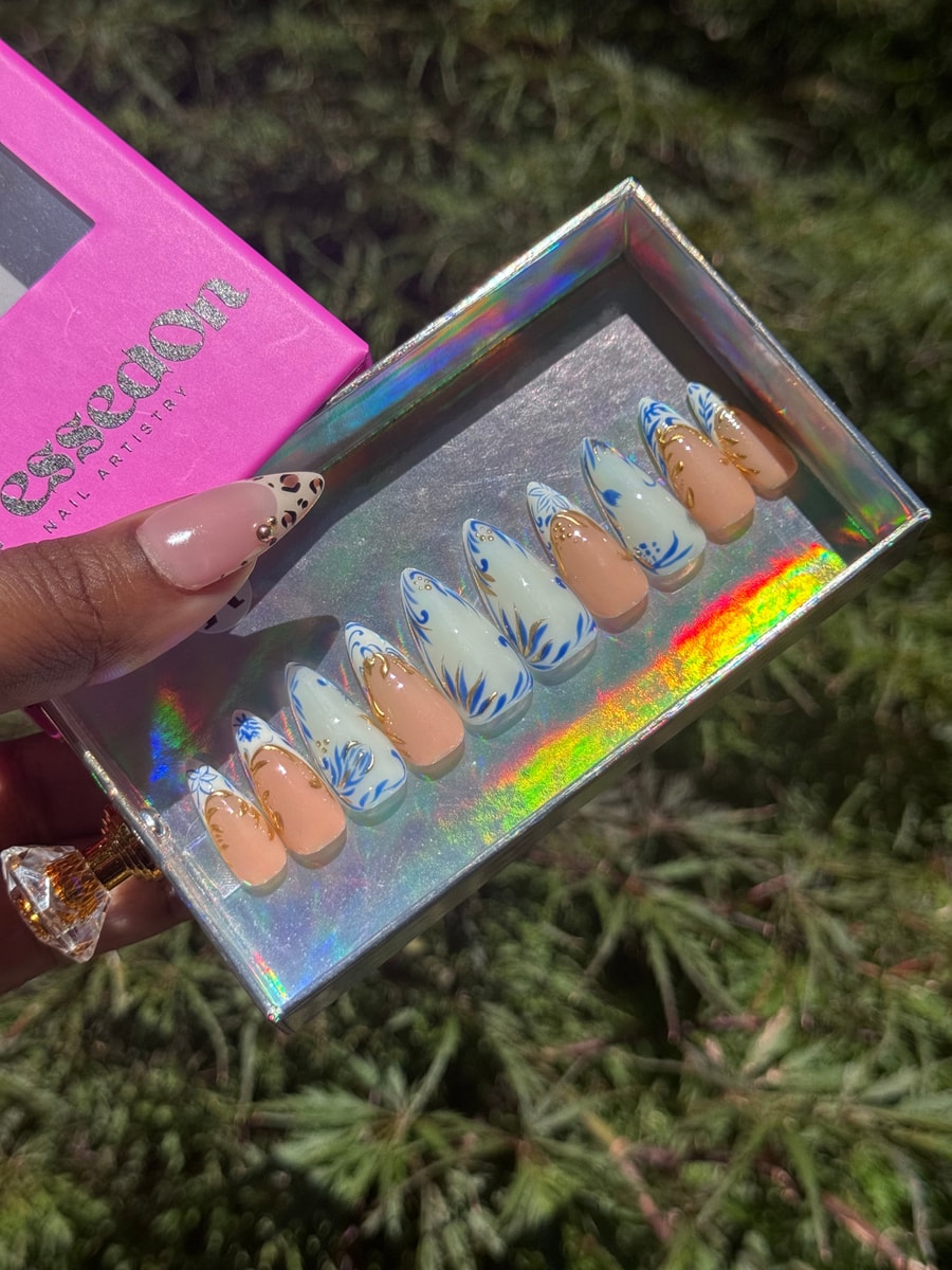 A set of short, almond-shaped press-on nails with a blue floral design. Peach and gold accents add detail to the white-based nails.