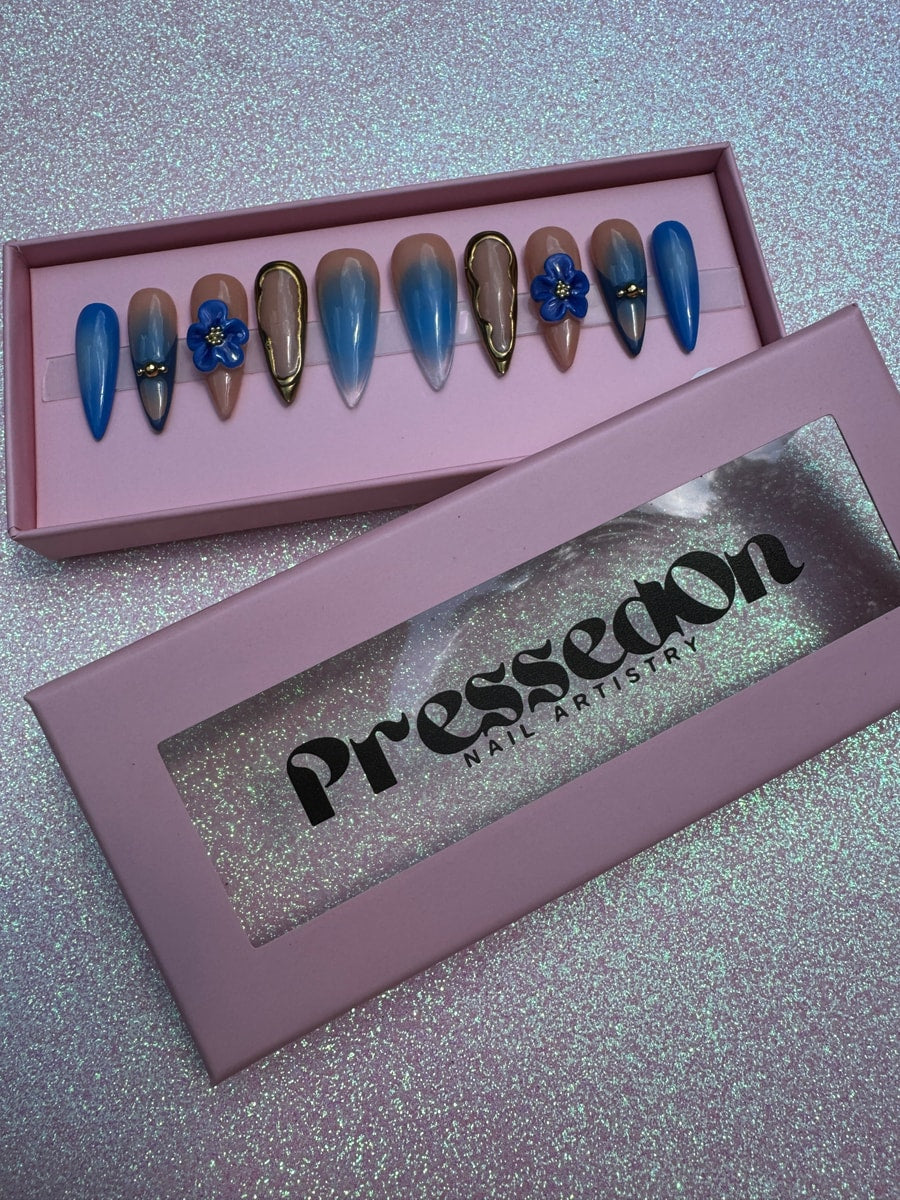 Medium-length press-on nails featuring royal blue and nude ombre design with gold accents. Some nails have 3D flower charms. The nails have a pointed shape.
