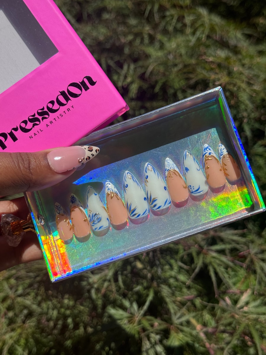 Short press-on nails with a blue and white floral design complemented by peach-colored nails with gold leaf accents.