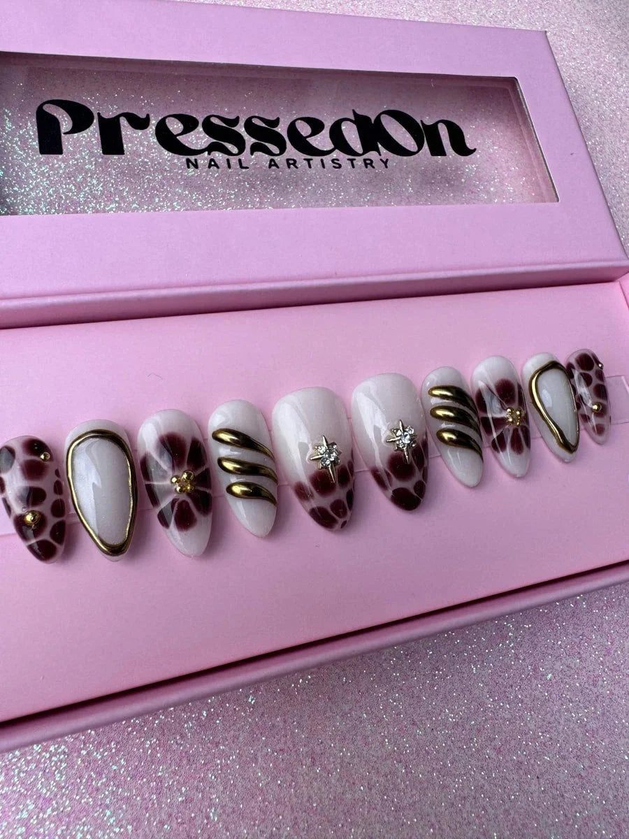 Super short press-on nails featuring bronzed brown, gold and white designs with floral patterns, geometric shapes, and spots, accented with shimmering stars and gold details.