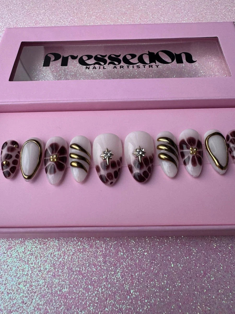 Super short press-on nails featuring bronzed brown and white designs, floral patterns, gold accents, and a star embellishment. The nails have a glossy finish.