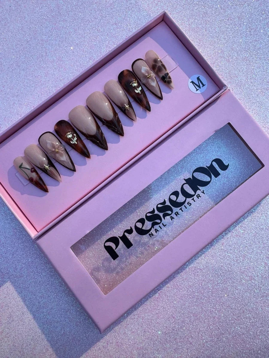 Set of medium brown stiletto press-on nails with a nude base and various designs including brown French tips, cherry, 3D flower, and gold star accents. Some nails feature a tortoiseshell pattern.