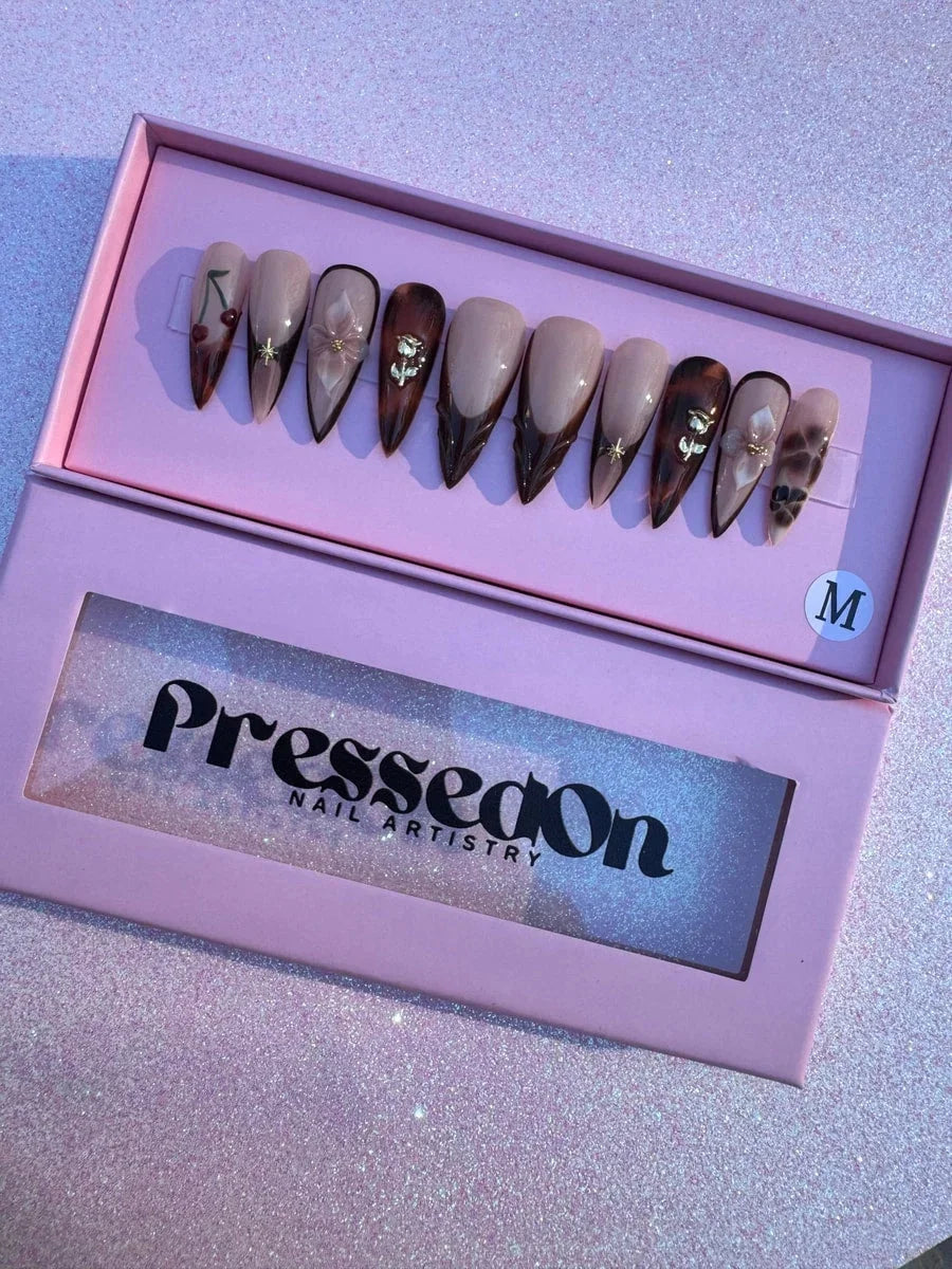 Medium length stiletto press-on nails in brown and nude tones with French tips. Designs include 3D flowers, 3D gold rose, gold stars, and cherry nail art.