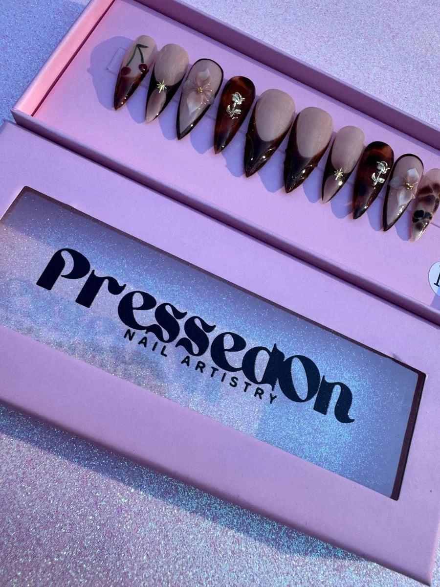 Medium length stiletto press-on nails feature brown and nude shades with French tips and floral accents. Nail art includes 3D gold roses and flowers, gold stars, and cherry designs, with some nails featuring a tortoiseshell pattern.