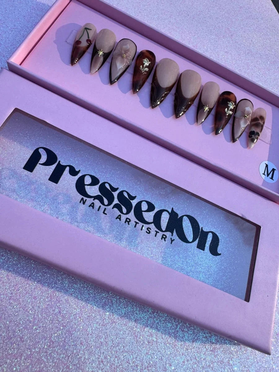 These are medium-length brown stiletto press-on nails with nude accents and designs including a cherry, 3D flower, and gold star charms. Some nails feature a brown French tip and tortoise shell pattern.