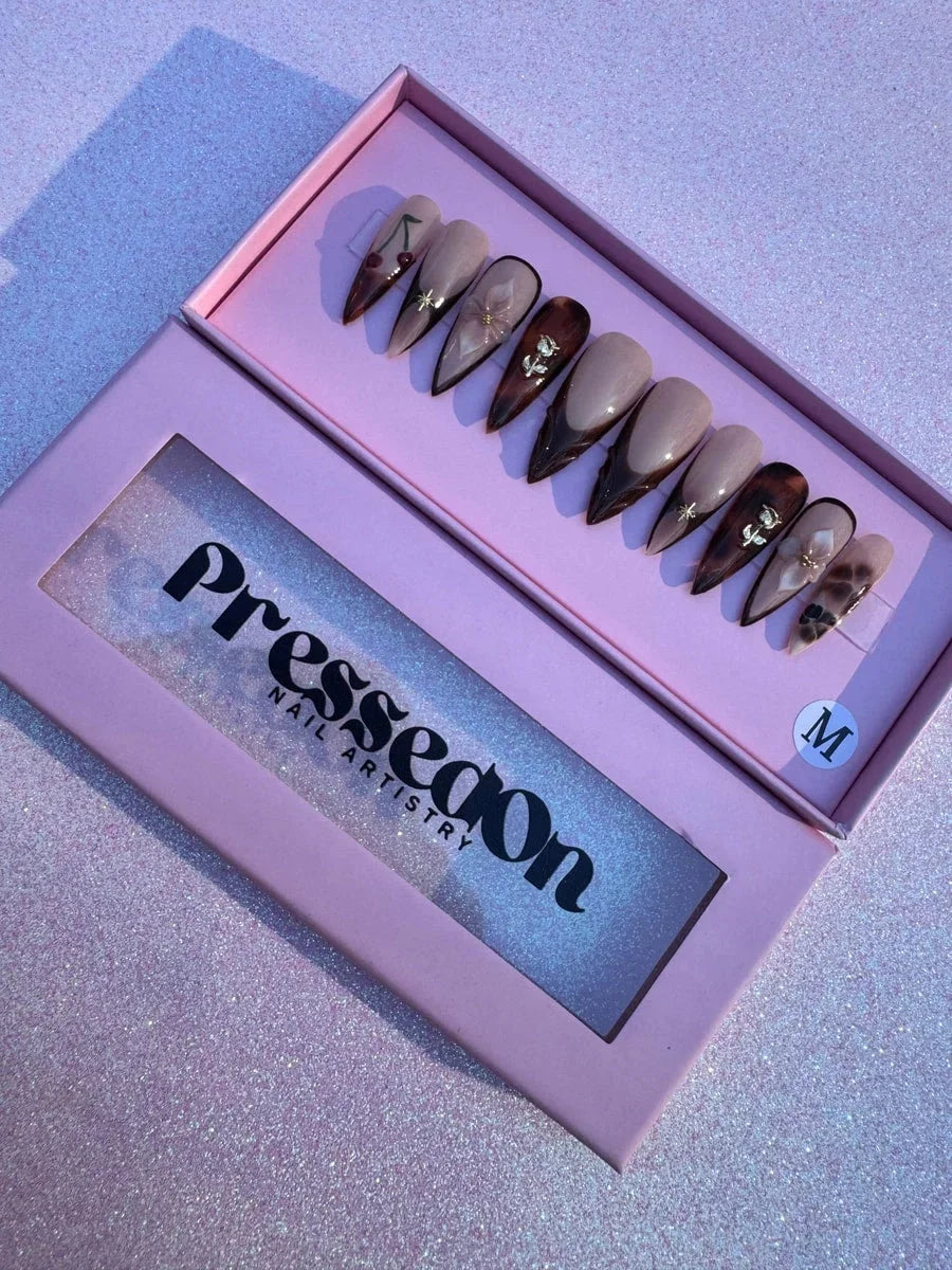 This is a set of medium-length brown stiletto press-on nails with nude bases, brown French tips, and floral, cherry, and star accents. The set features 3D flowers and roses, and gold star charms.
