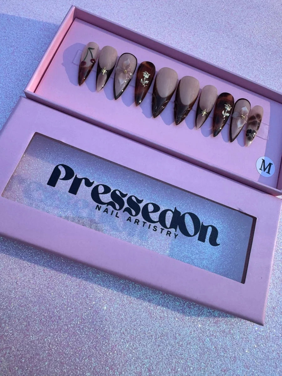 Medium length brown stiletto press-on nails feature nude bases, brown French tips, a cherry design, 3D gold rose, 3D flower, gold star accents, and tortoise shell detailing.