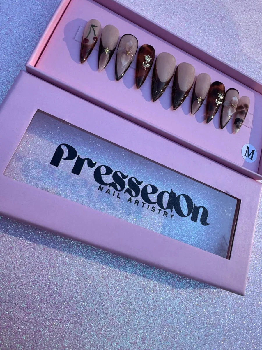 These are medium-length brown stiletto press-on nails with a nude base. The designs include brown French tips, tortoise shell patterns, as well as cherry, 3D flower, gold star and gold rose accents.