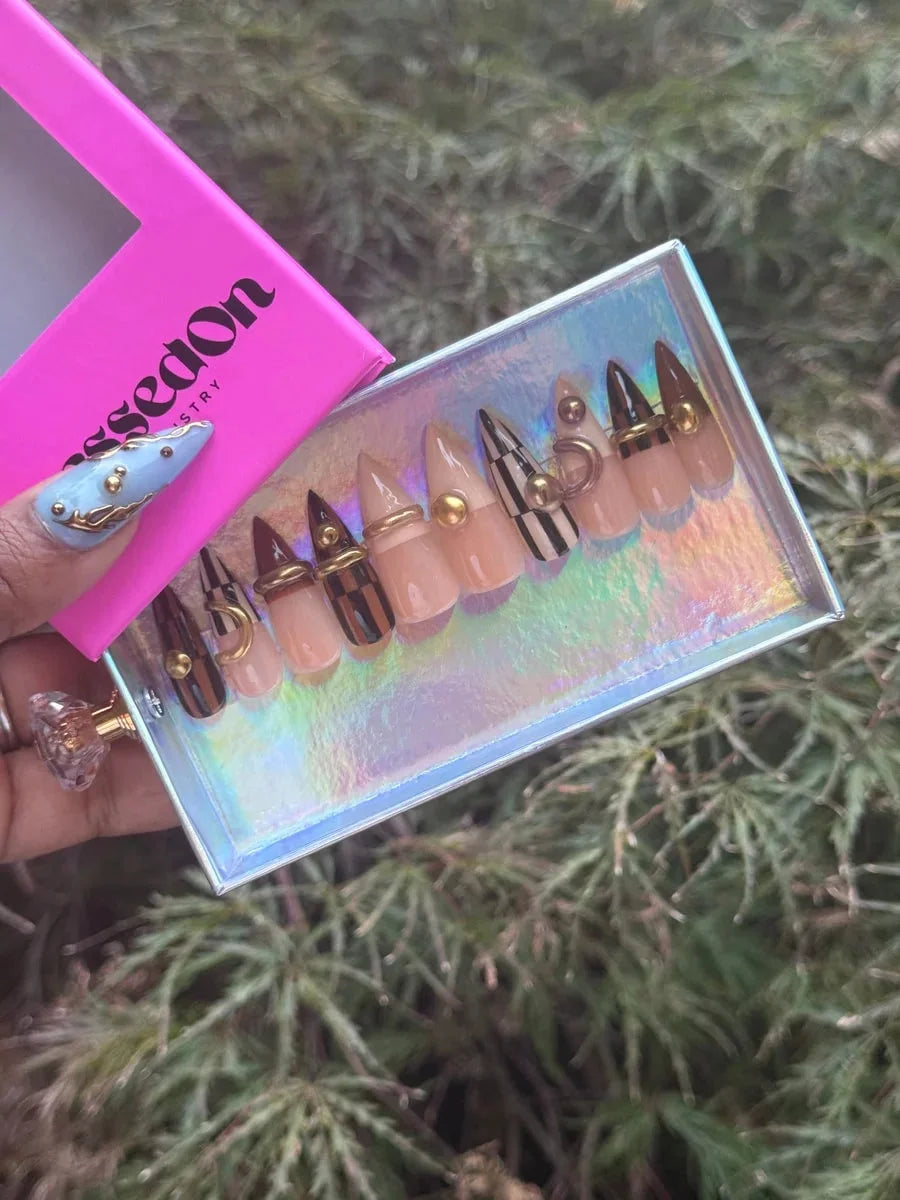 Set of stiletto press-on nails featuring brown, tan, and checkered designs, accented with gold rings and studs. The nails have nude bases with brown tips and gold embellishments.