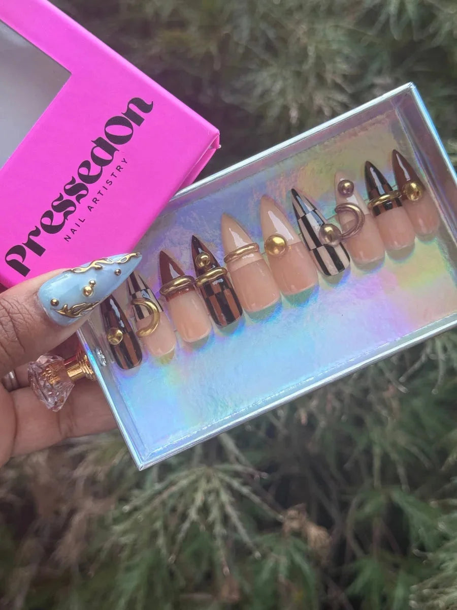 Short stiletto press-on nails featuring brown, tan, and nude shades, with checkerboard designs, gold studs and rings. Some nails feature brown tips and nude bases.