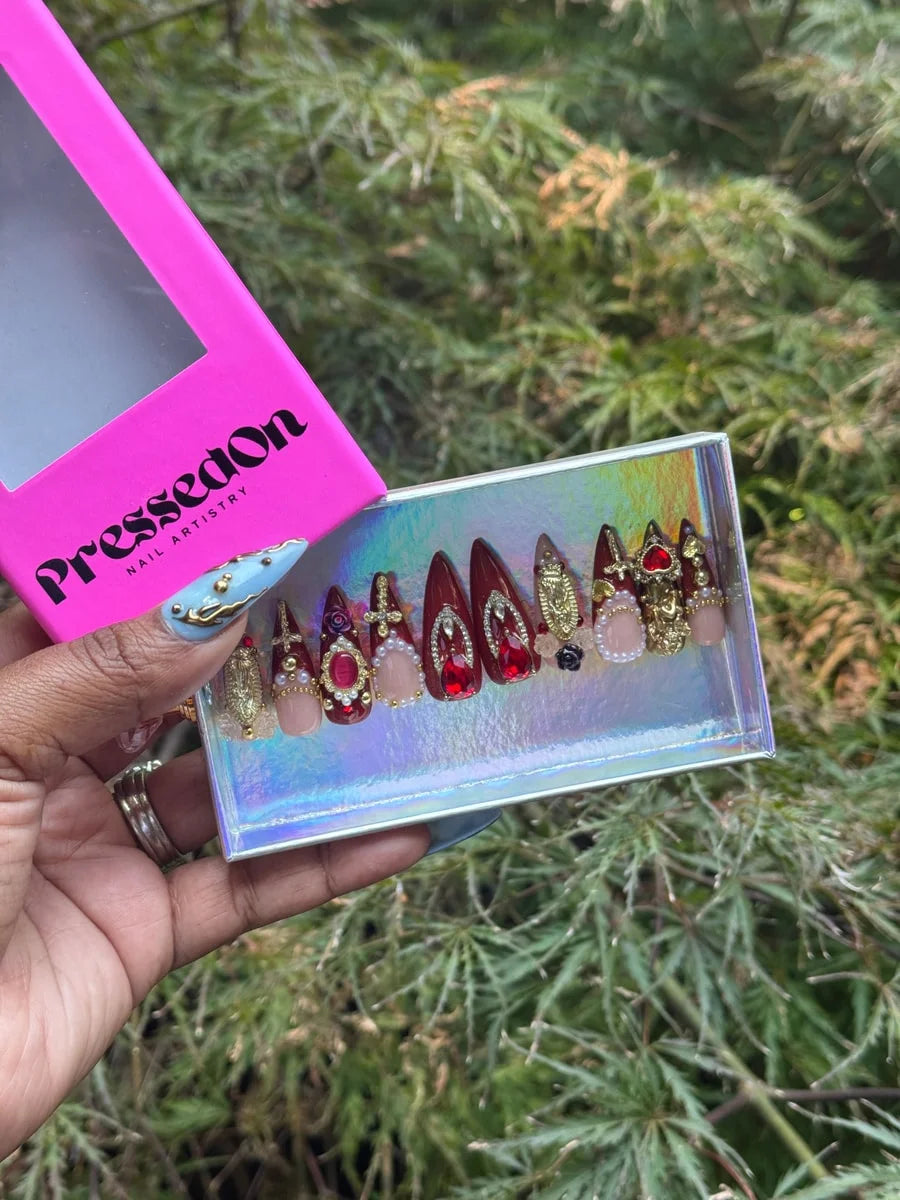 Set of candy apple red stiletto press-on nails decorated with pearls, rhinestones, crosses, hearts, and gold accents. Some nails are nude with matching embellishments.
