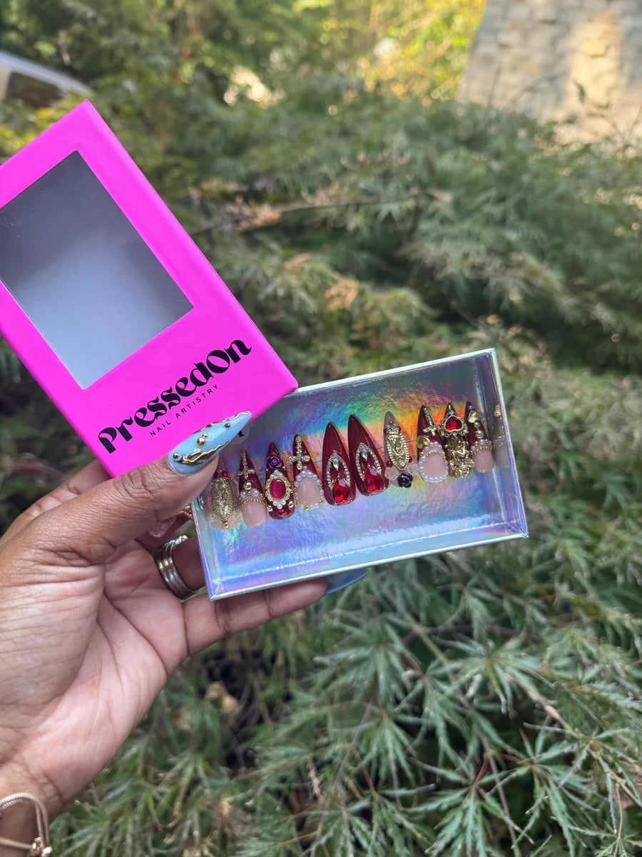 Candy apple red short press-on nails adorned with gold hearts, crosses, pearls, rhinestones, and charms. Includes nude accent nails with pearl embellishments.