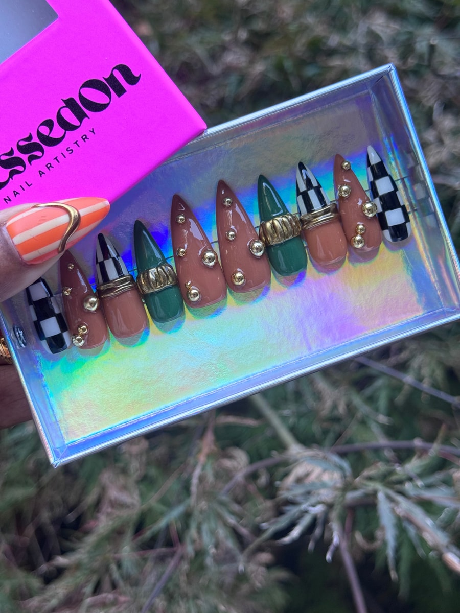 Medium stiletto press-on nails featuring brown, green, and checkerboard patterns with gold ring and stud embellishments on the tips.