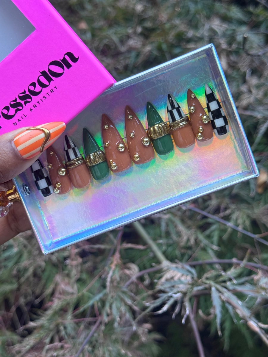 Medium stiletto press-on nails include checkmate, green, and brown designs. Gold studs and ring embellishments add detail to some of the nails.
