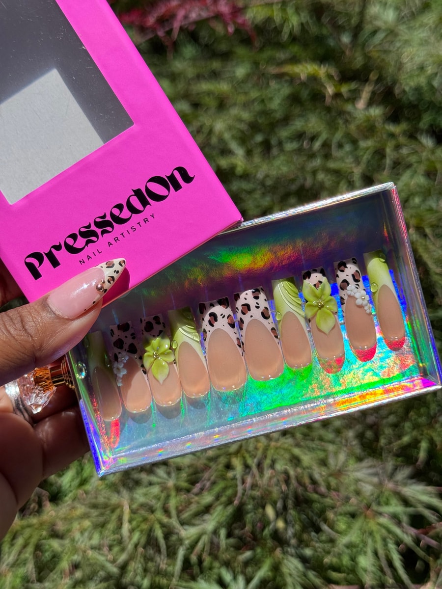 A set of medium-length press-on nails with cheetah print, lime green tips, and floral accents. One nail has white flowers, and another has a neon pink tip.