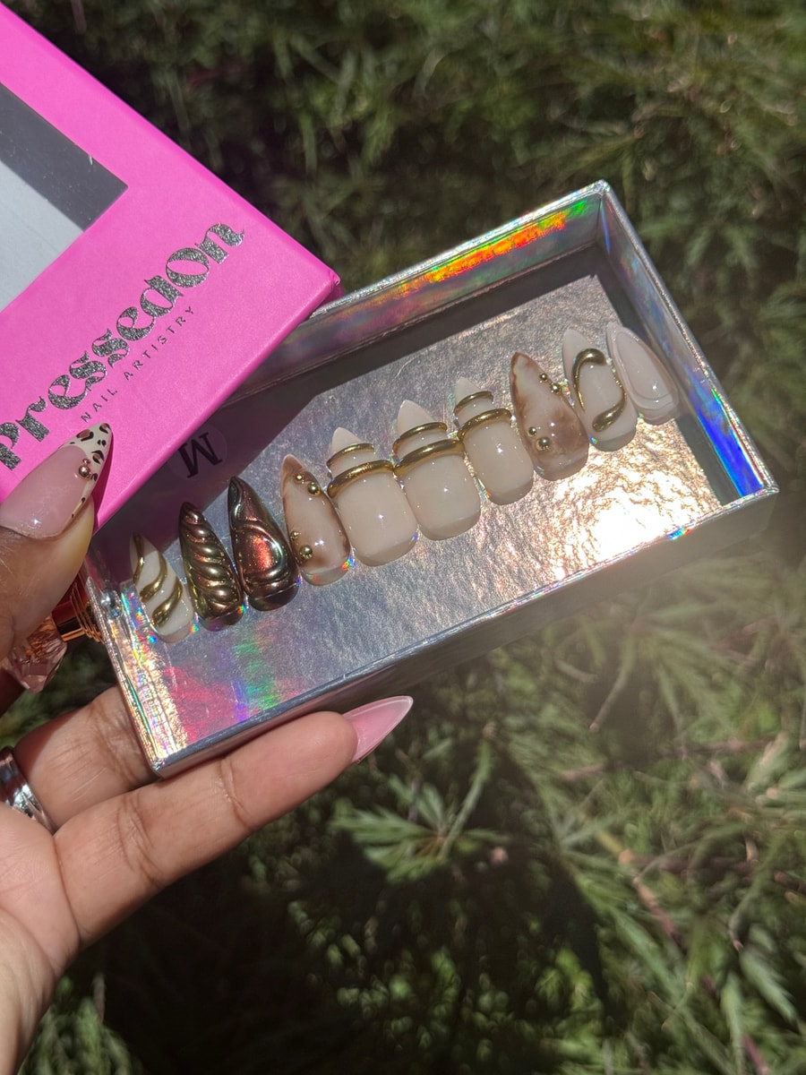 A collection of short press-on nails with chrome, gold bead, nude, brown, and white designs. Several nails feature gold swirls or bands.