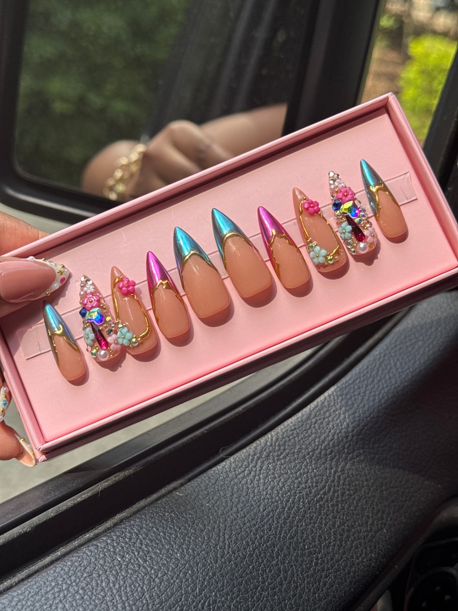 Set of nude medium-length press-on nails with gold accents, iridescent chrome French tips, gem stones, and floral decorations.