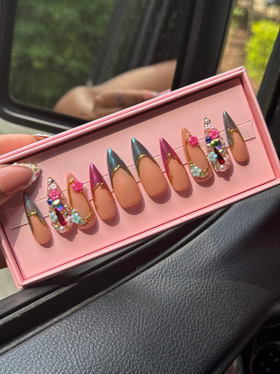 A set of medium almond-shaped press-on nails with nude bases, chrome and gold french tips, and decorative gems and flower charms. Some nails have pink or iridescent chrome tips.