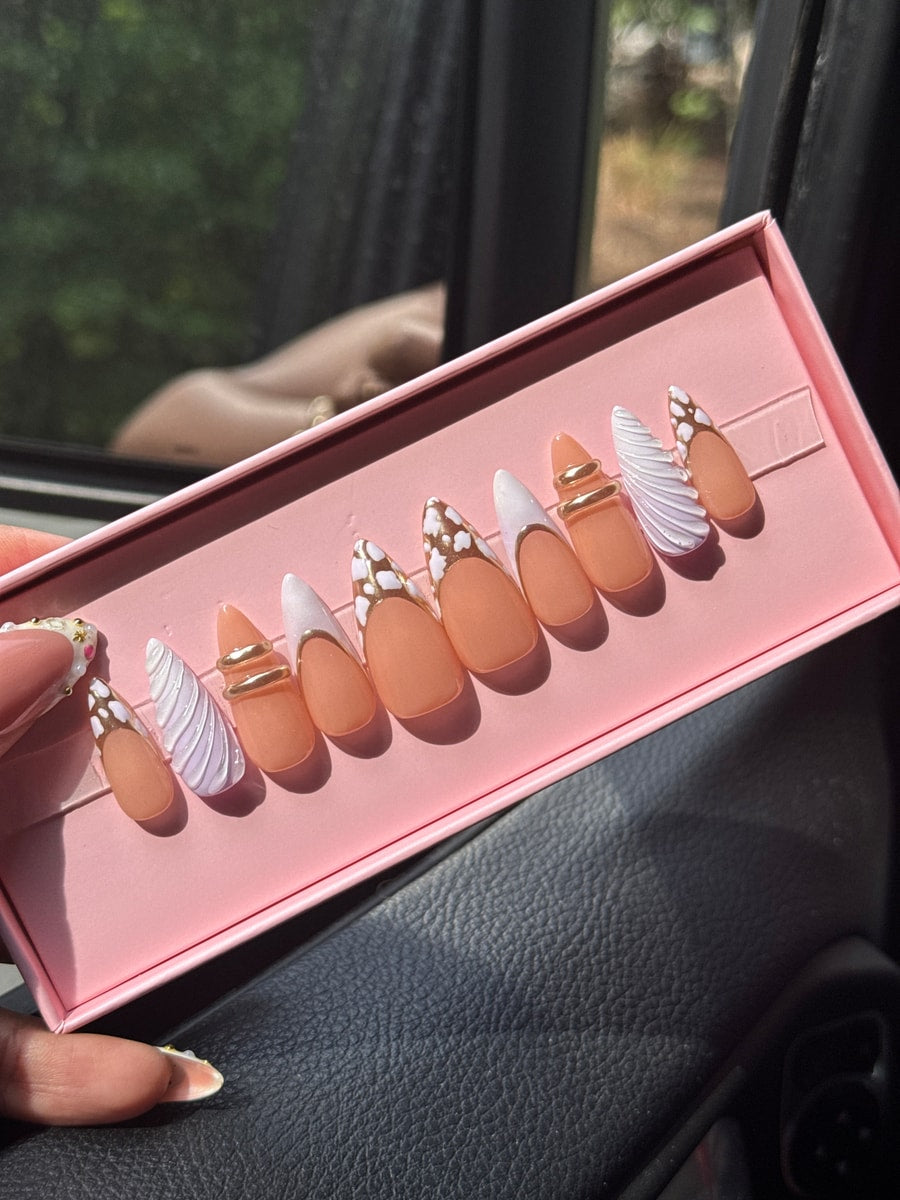 Assorted press-on nails with a chrome and cloud-like finish, featuring white accents, swirling details, gold rings, and nude bases with patterned tips.