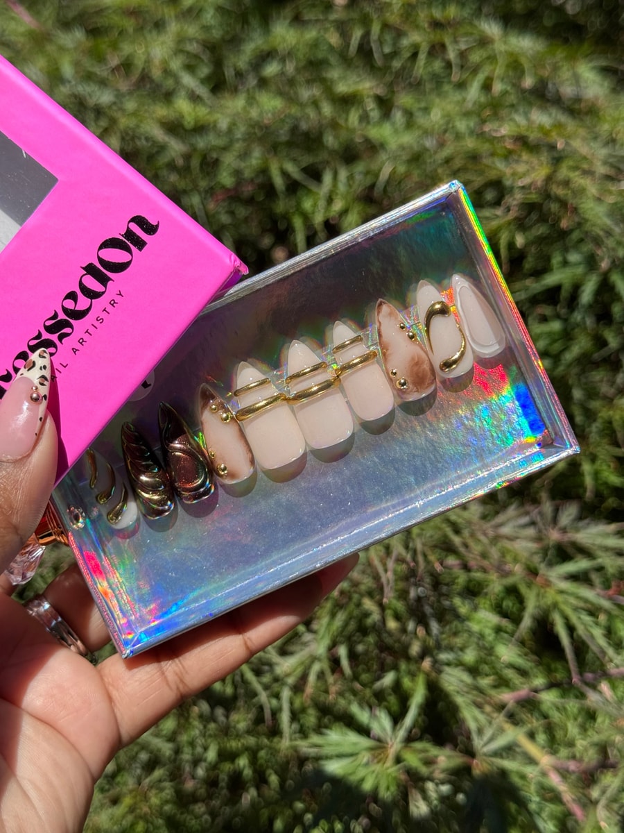 Short neutral press-on nails with chrome accents, gold rings, marbled patterns, and small gold embellishments. Some nails are fully metallic chrome.