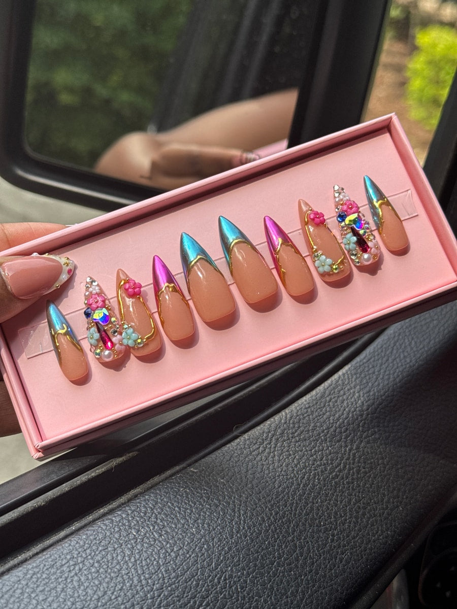 This is a set of medium press-on nails with a nude base. Some nails have chrome tips and gold accents; others feature gems, rhinestones, and 3D flower decorations.