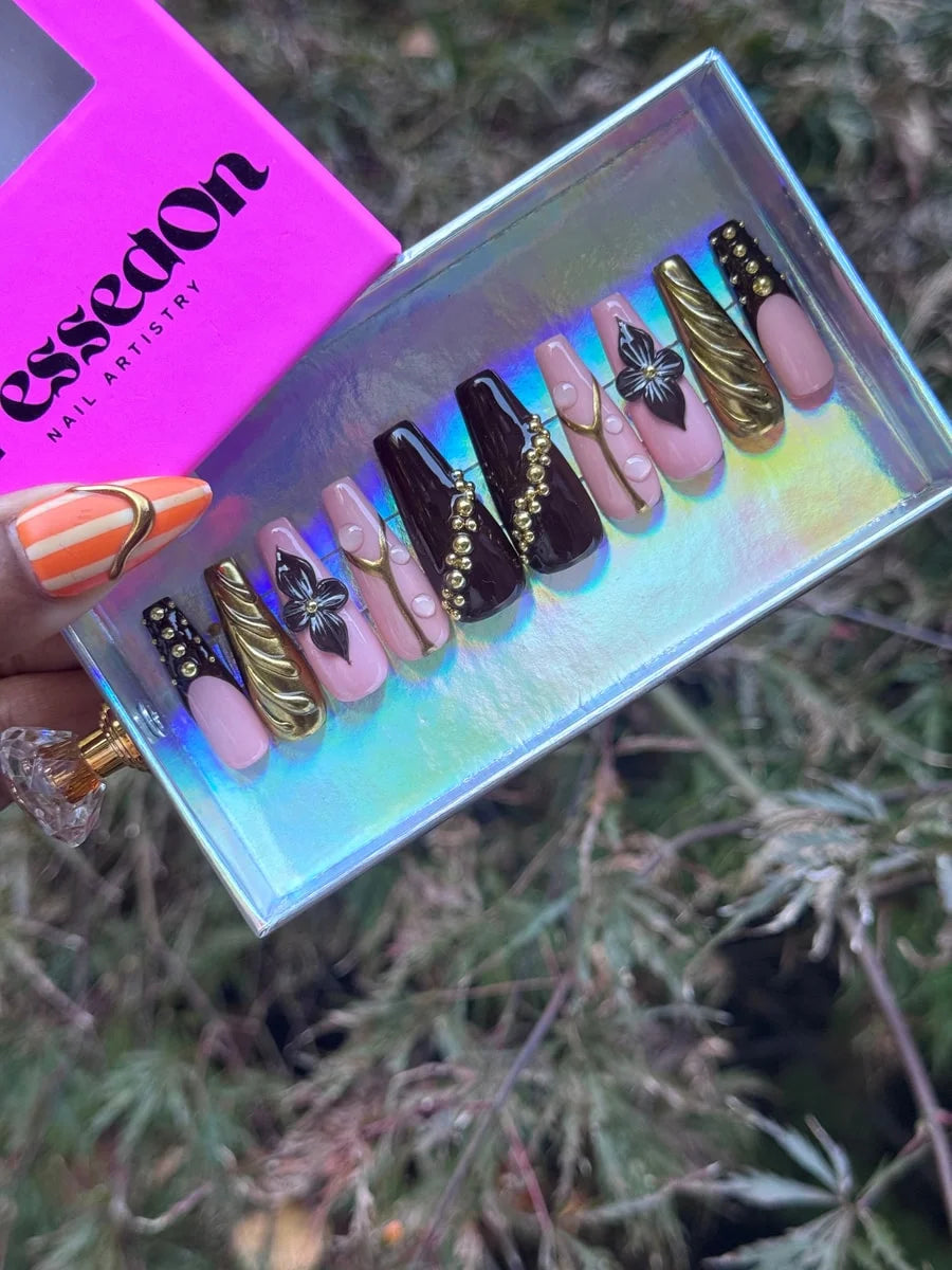 A set of cocoa brown and light pink press-on nails with gold details. The nails feature various embellishments, including gold tips, beaded accents, and floral designs.