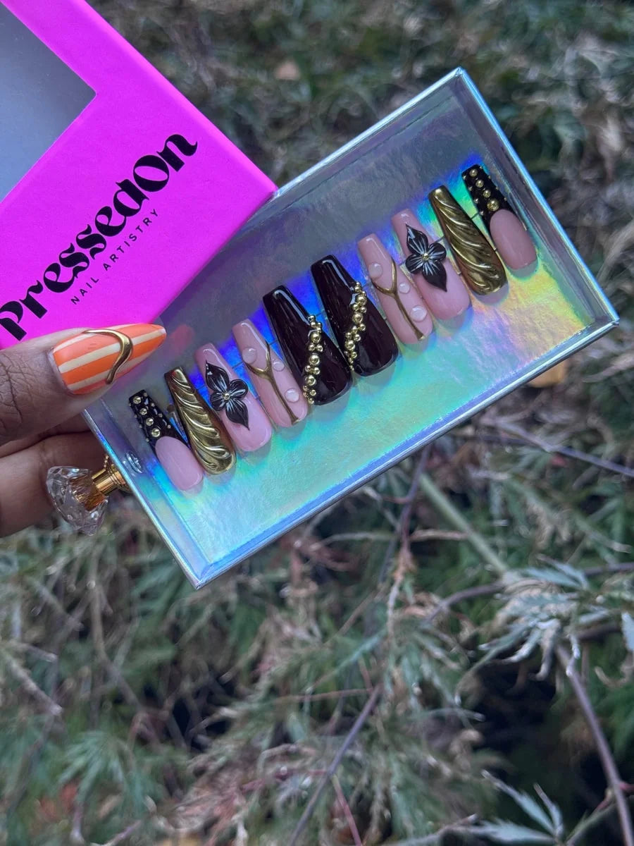 A set of cocoa luxe press-on nails in brown and pink tones are displayed. The nails are medium length and feature gold embellishments, floral designs, and french tips.