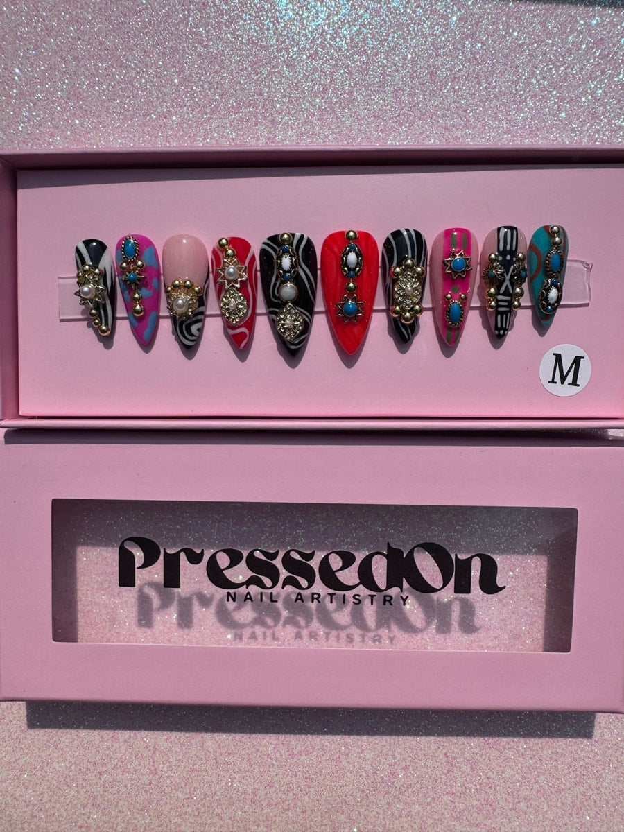 A set of short, stiletto press-on nails with colorful patterns and charms including pearls, turquoise, and gold accents. The nails are arranged in a display box.