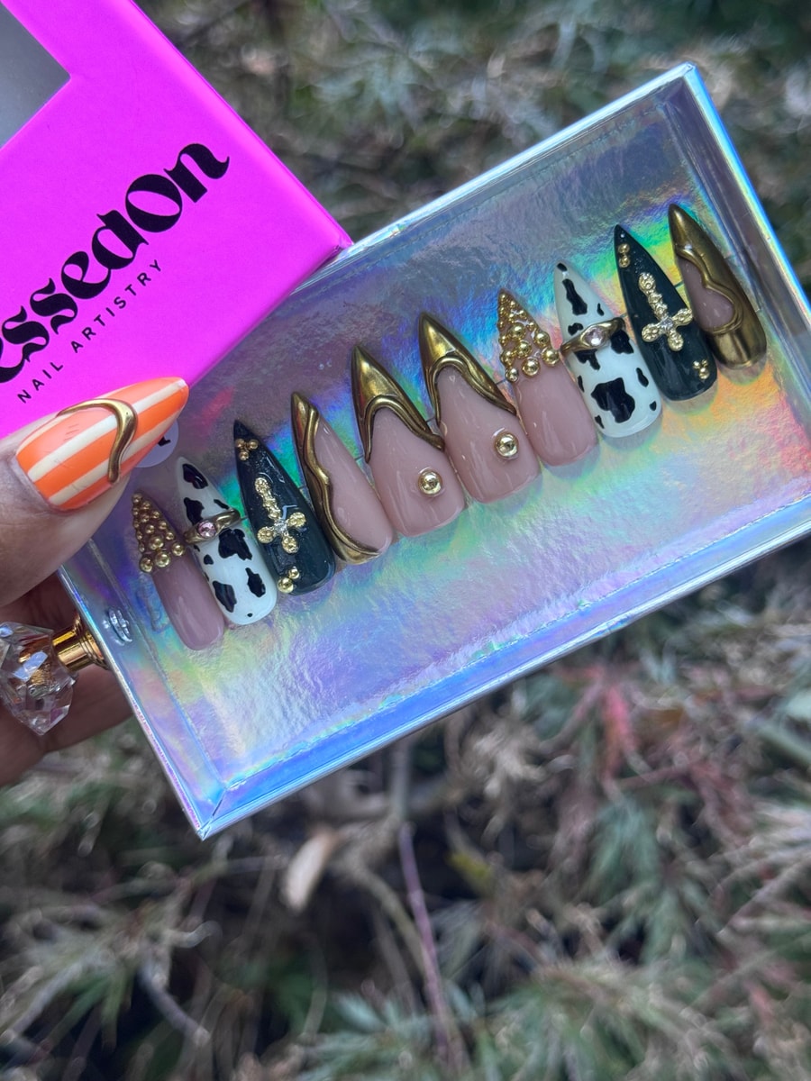 These press-on nails feature a "Cowgirl Couture" style with designs including cow print, crosses, rhinestones, and gold detailing on pink, black, and white nails.