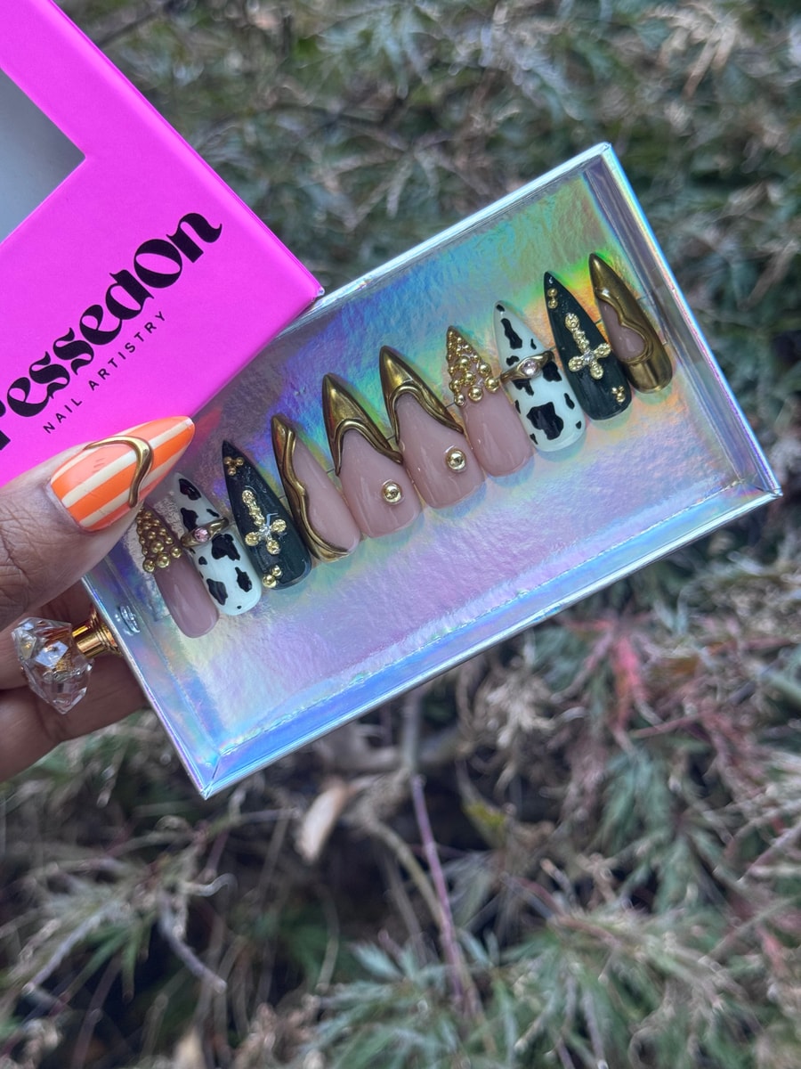 Medium length press-on nails feature cow print, gold details, crosses, and nude shades for a unique cowgirl style.