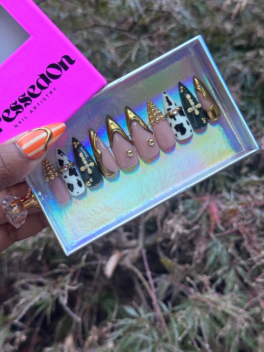 Set of medium-length press-on nails in a cowgirl couture style. The nails feature cow print, gold detailing, studded embellishments, and a color palette of nude, black, and gold.