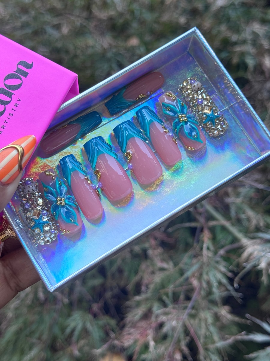 A set of medium-length pink press-on nails with a teal tide design and gold embellishments. One nail has a 3D teal flower, and another is covered in rhinestones.
