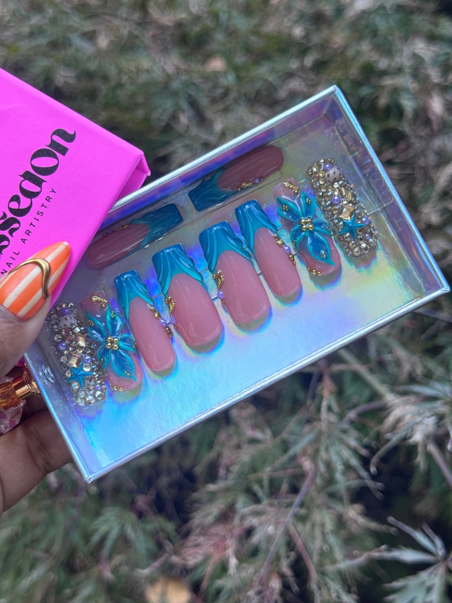 Medium-length pink press-on nails with teal French tips featuring wave-like accents, rhinestones, gold embellishments, and 3D flower charms.