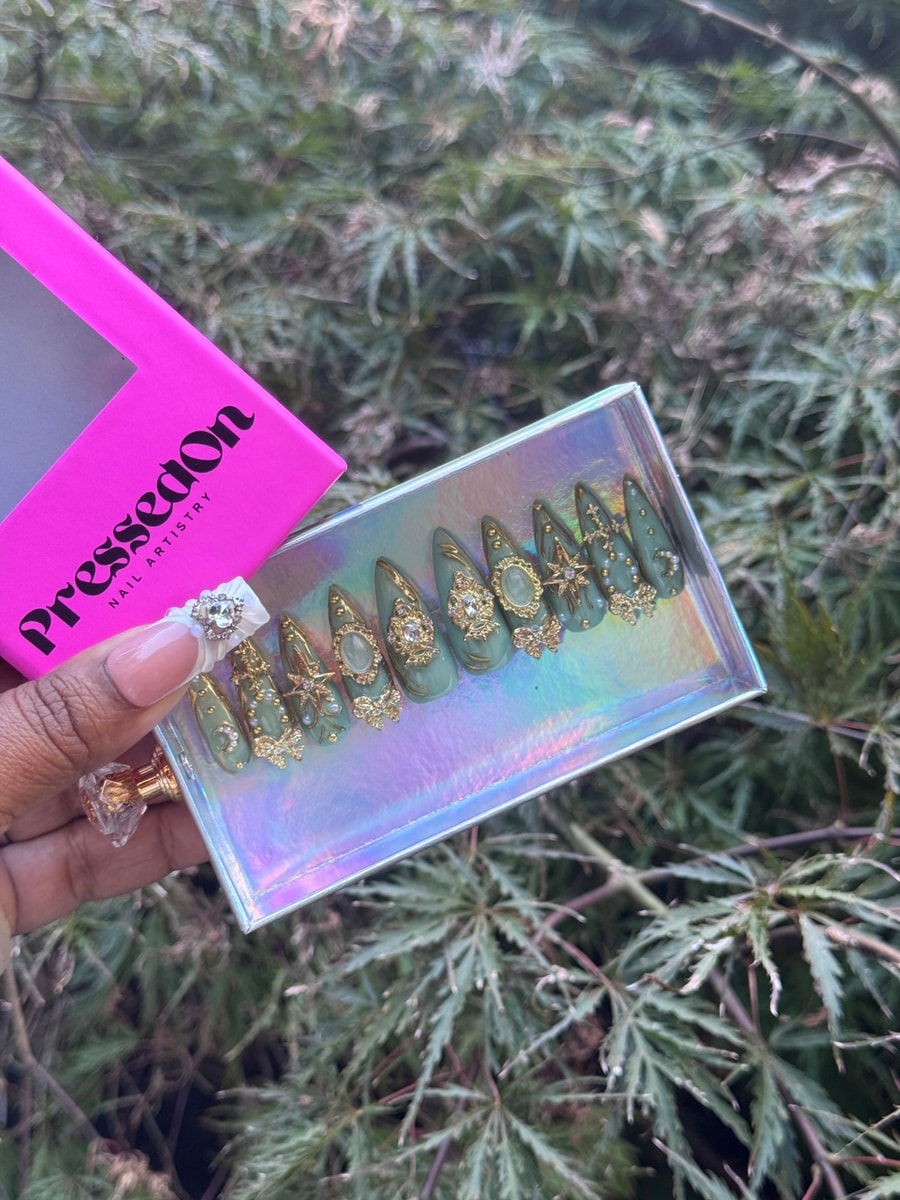 Emerald green press-on nails with medium length and gold embellishments. The nails feature various designs including starbursts, moons, gemstones, and beads.