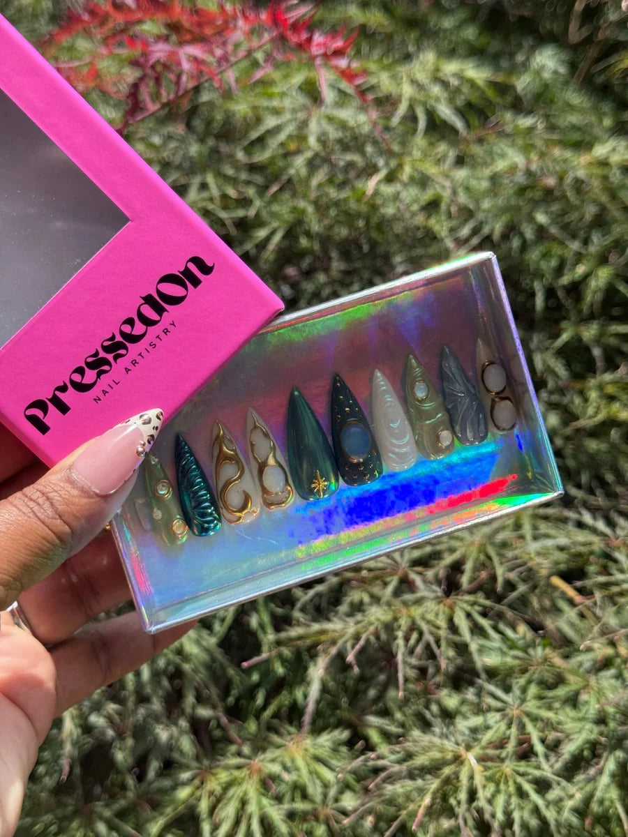 Medium-length press-on nails with charmed, celestial designs featuring green, gold, white, and blue colors with gemstone and star accents. The nails showcase moonstone details and orb embellishments.