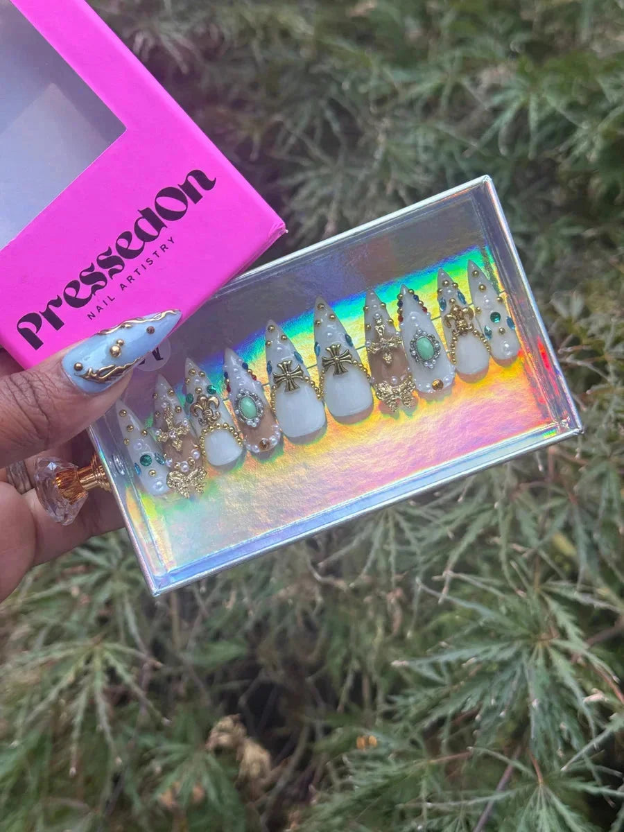 These are short stiletto press-on nails in a white and nude palette, decorated with Fleur de Lis, Iron Cross, pearls, gold embellishments, and gem accents.