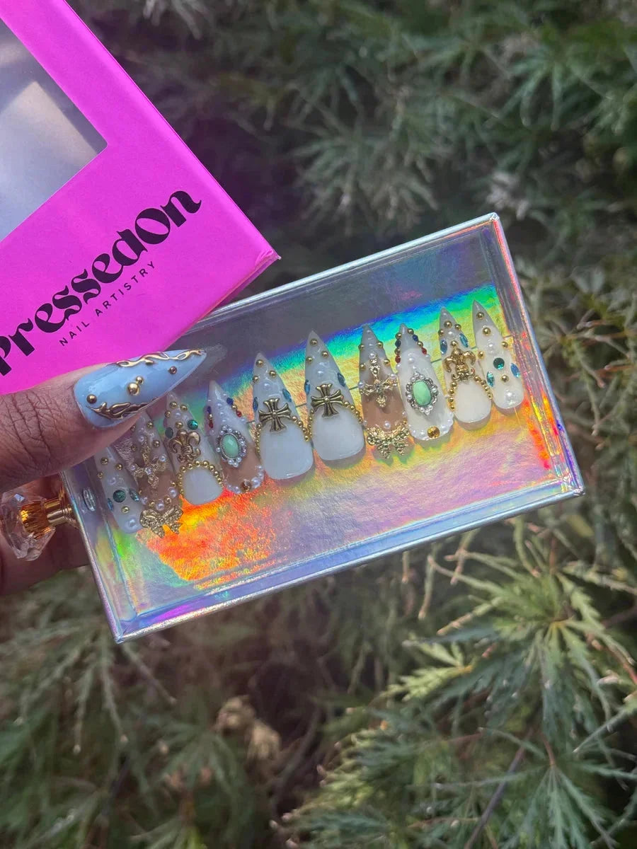 Short stiletto press-on nails in a white and nude color scheme, adorned with gold Fleur de Lis and Iron Cross accents, pearl beads, and gem details.