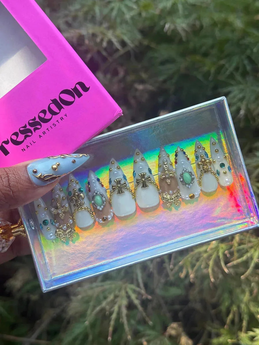 Short stiletto press-on nails with a white base, adorned with gold fleur de lis, iron cross, pearl, and gem embellishments.