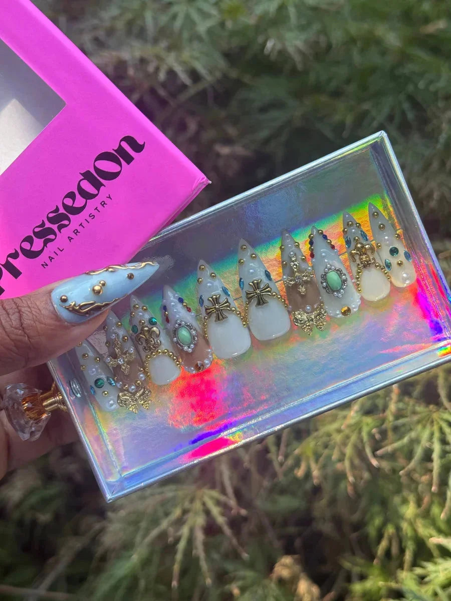 Short stiletto press-on nails in white and nude, adorned with gold fleur de lis, iron cross embellishments, and pearl gems. The design creates an angelic and elegant look.