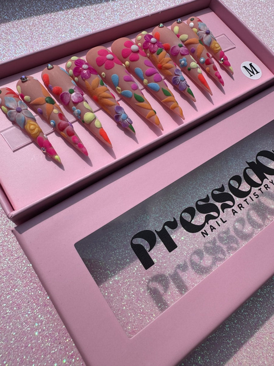 Set of extra long press-on nails with a nude base and colorful floral designs. Each nail has 3D flower accents in pink, orange, blue, and green, with some nails also having small gem accents.