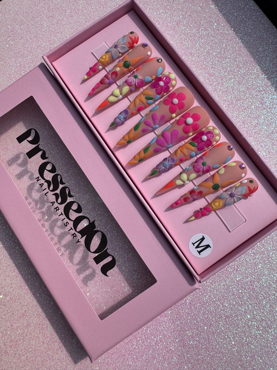 Extra long press-on nails featuring a nude base adorned with colorful 3D floral designs in pink, purple, orange, and yellow, accented with gems.