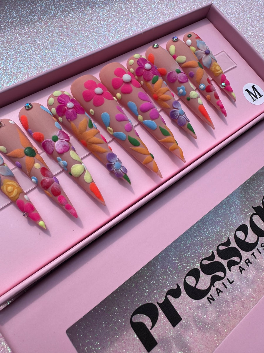 These are extra long stiletto press-on nails with a nude base, adorned with colorful 3D floral embellishments.