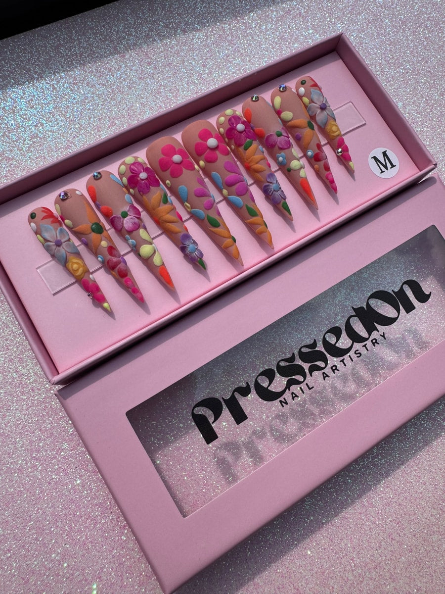 These are stiletto-shaped press-on nails with a nude base. Each nail is decorated with a variety of colorful, three-dimensional flowers. Some nails have rhinestone accents.