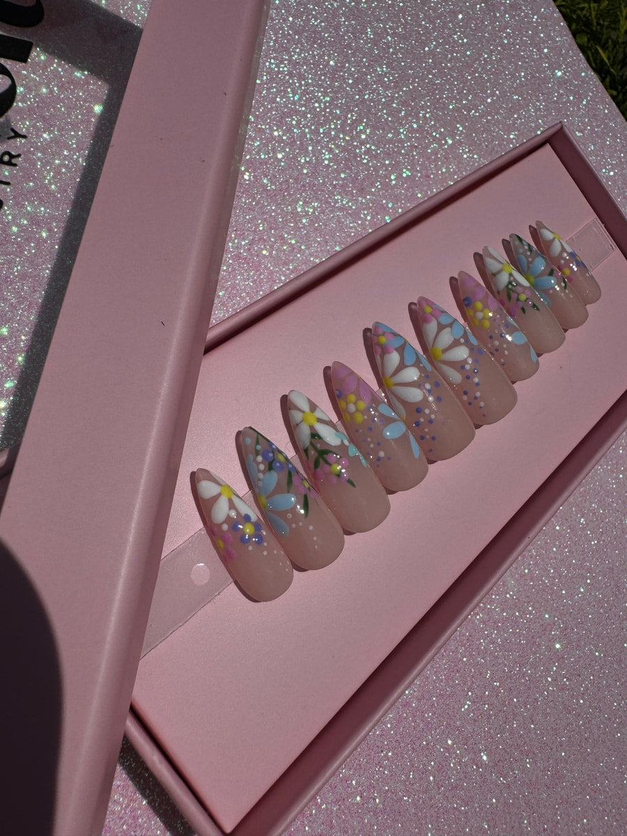 Short almond-shaped press-on nails with a nude base, adorned with a floral garden design. Features white daisies with blue and pink accents, and small dot embellishments.